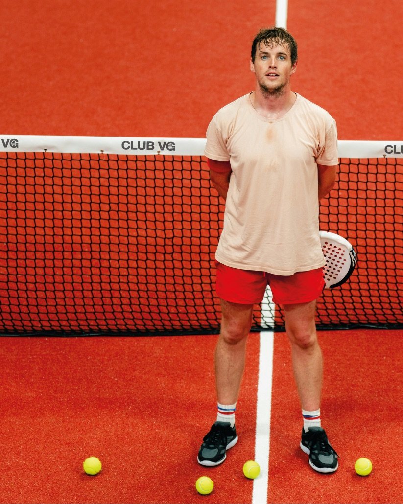 Joueur de padel devant un filet siglé Club VG, sur un terrain rouge.